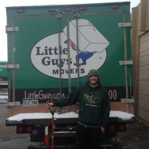Mover holding a shovel and standing next to Little Guys Movers truck while snow falls