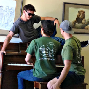 Two movers playing piano with one mover in sunglasses sitting on top of piano