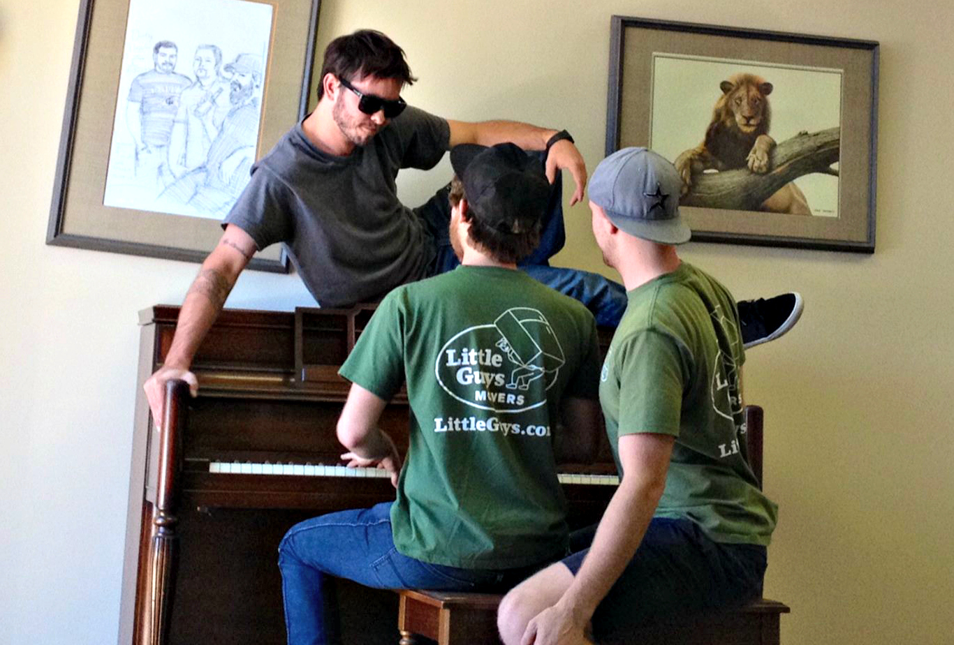 Two movers playing piano with one mover in sunglasses sitting on top of piano