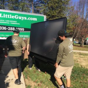 Two movers carrying a flatscreen TV to a Little Guys Movers truck