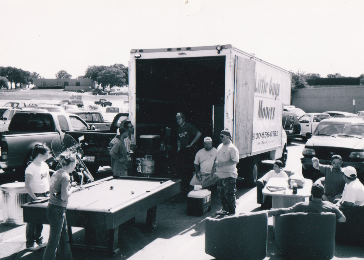 An early black-and-white photo of a group of Little Guys gathered around a pool table near a moving truck