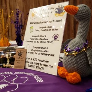 a table at Mardi Paws 2019 decorated with purple table cloth, a stuffed animal goose, and the night's schedule