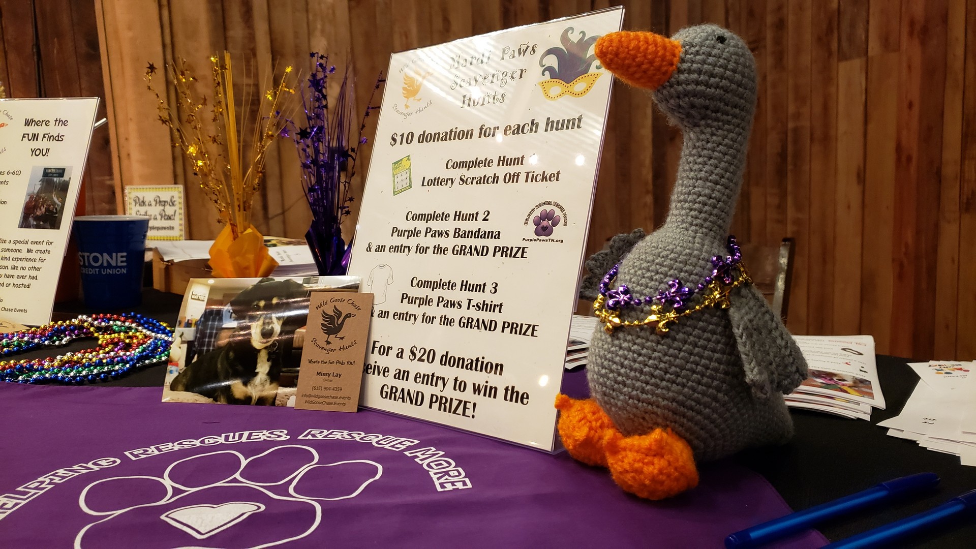 a table at Mardi Paws 2019 decorated with purple table cloth, a stuffed animal goose, and the night's schedule