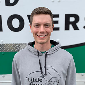 Little Guy Alex wearing a grey Little Guys Movers hoodie and smiling in front of one of the moving trucks