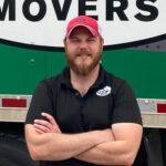 A Little Guys Movers manager wearing a black polo shirt (and Little Guys Movers logo) and red hat stands in front of a green moving truck, arms folded and smiling warmly