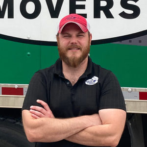 A Little Guys Movers manager wearing a black polo shirt (and Little Guys Movers logo) and red hat stands in front of a green moving truck, arms folded and smiling warmly