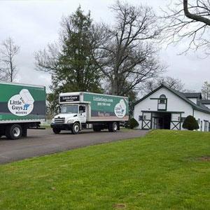 Two green Little Guys Movers moving trucks roll dreamily over a small road between bright green hills in Lexington