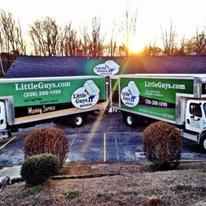 Two Little Guys Movers moving trucks are parked awesomely in front of the office; a ray of morning sunlight glows triumphantly behind them