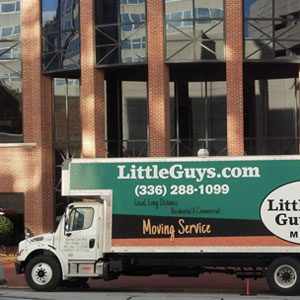 A Little Guys Movers moving truck is parked in front of a glass-and-brick condominium lobby entry.