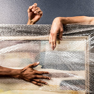 Hands from two Little Guys wrap a framed artwork in bubble wrap.