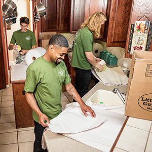 Three professional Little Guys Movers wrap kitchen items in paper and pack them into moving boxes