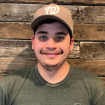 A Little Guys Movers mover stands in front of a wooden plank background, wearing a tan ball cap.
