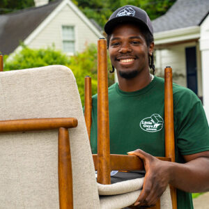 A Little Guys Movers mover, wearing a hat and green shirt, moves furniture to the moving truck on a crisp fall day.