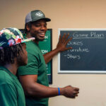 A Little Guys Movers mover points to a "Game Plan" written on a blackboard. Several other Little Guys Movers movers listen attentively.