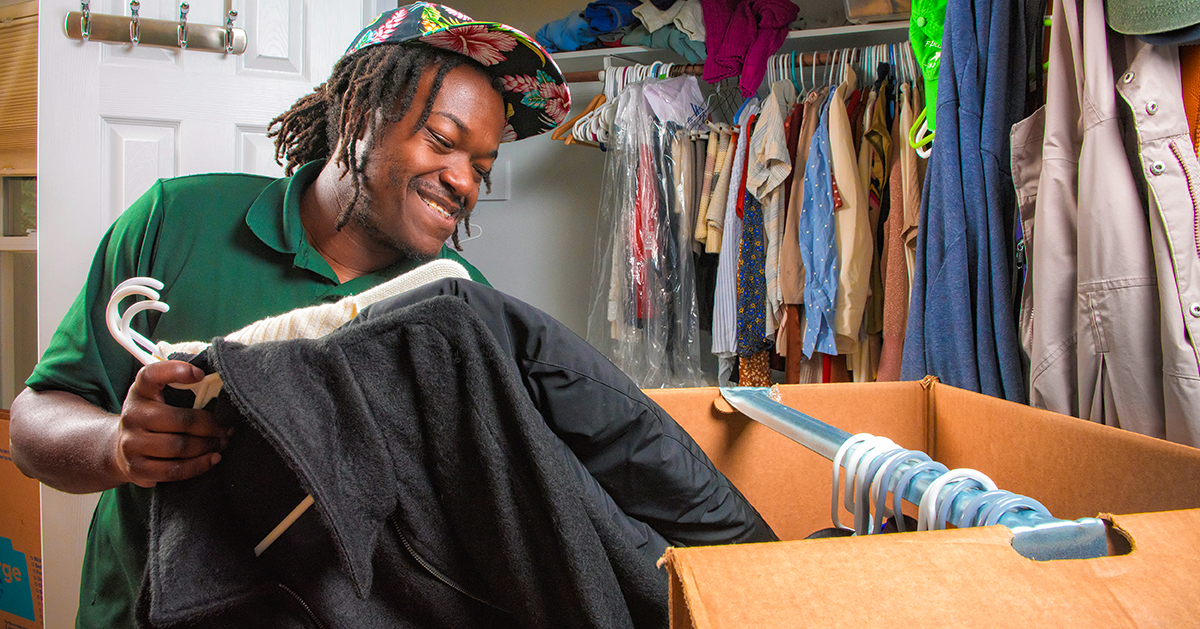 A Little Guys Movers mover smiles as he carefully packs clothes into a moving box.